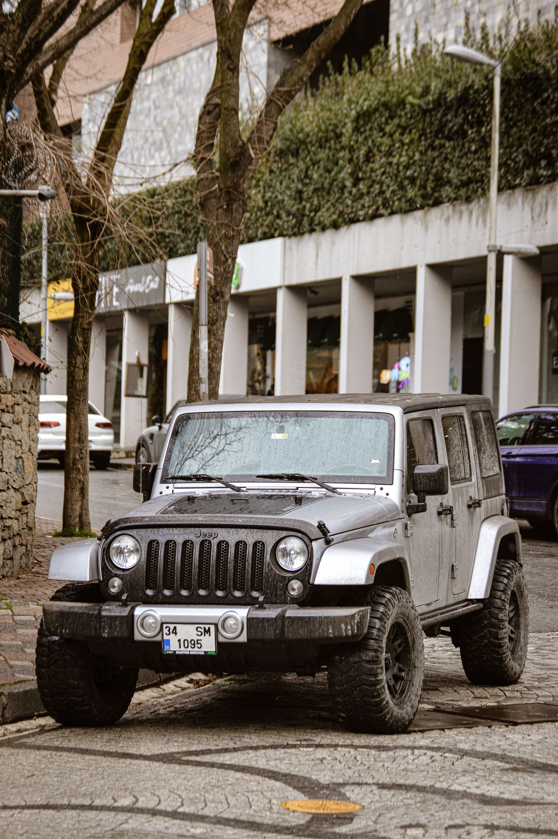 A rugged Jeep Wrangler parked on a city street surrounded by trees and buildings.